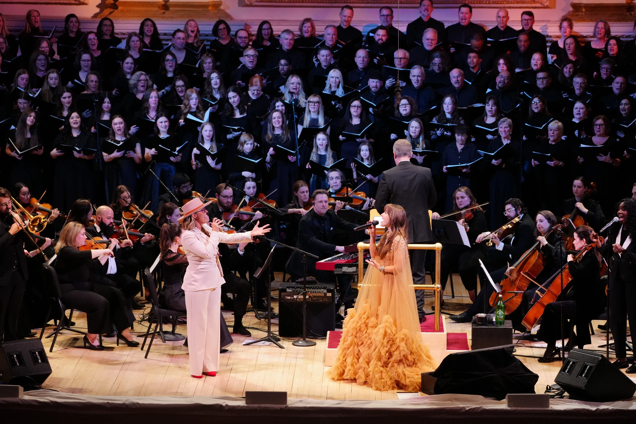 Two women sing onstage with an orchestra and large choir behind them in a concert hall, showcasing the transformative power of music therapy in patient care.