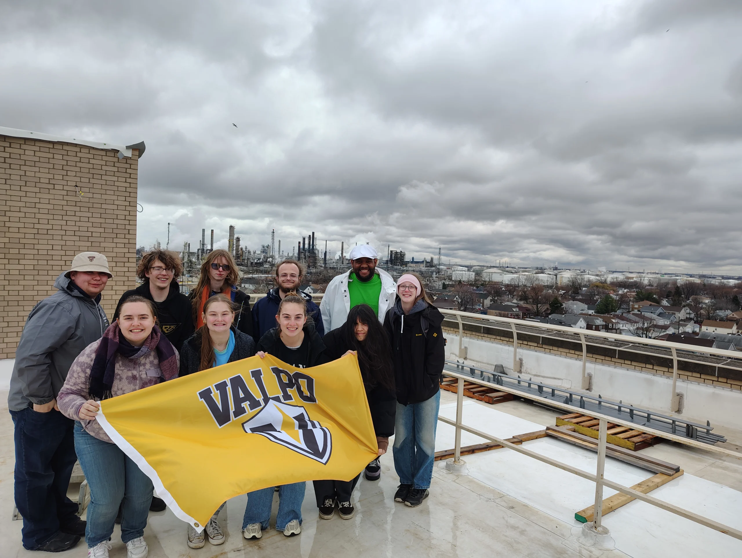 A group of Valparaiso University students serve together, holding a Valpo flag on a rooftop with an industrial cityscape in the background during spring break 2026.