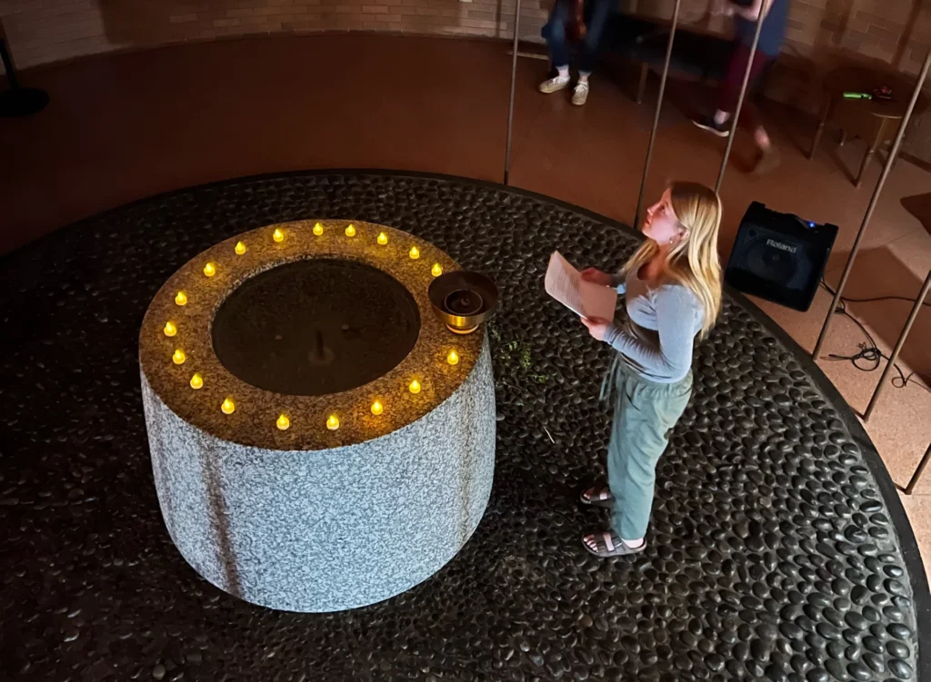 A woman speaks by a round stone altar with candles, as people sit on upper and lower levels of a circular room, evoking the contemplative atmosphere of Religious Studies at Valparaiso University.