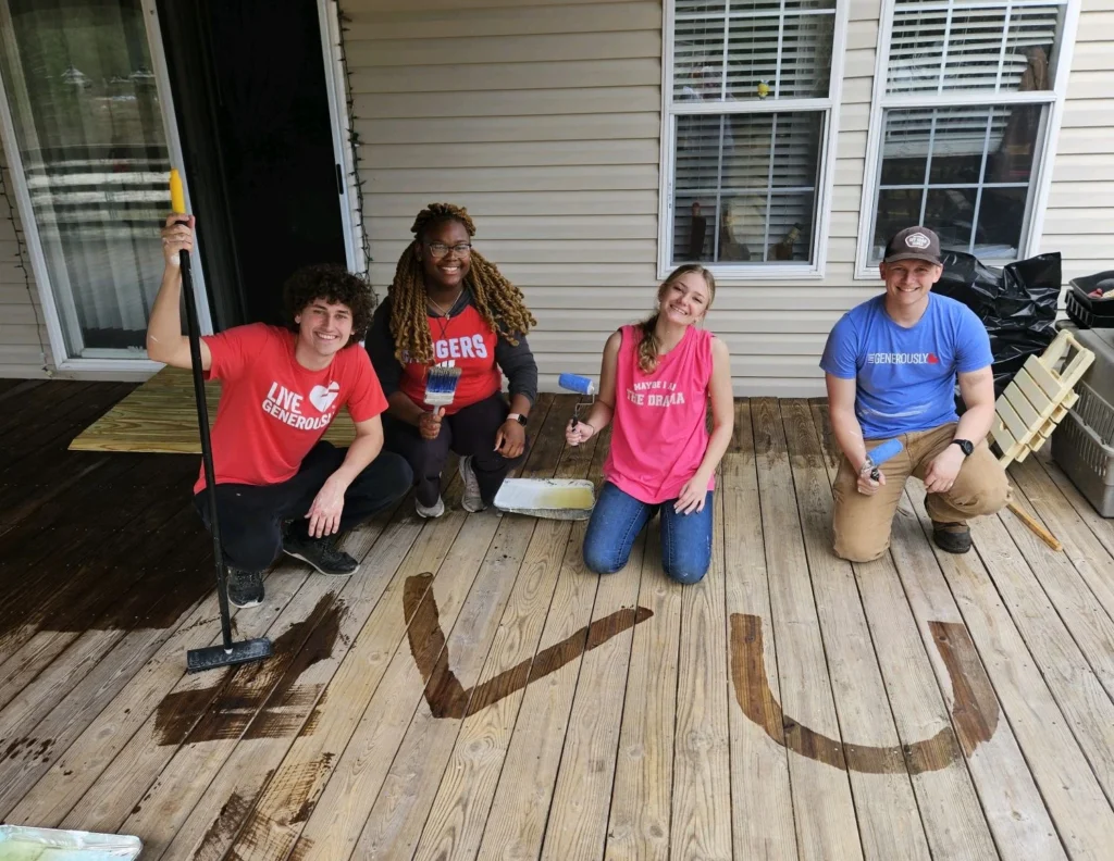 Four Valparaiso University students kneel and smile on a deck with "EVU" stained on the wooden boards. Deck supplies are nearby as the students serve during spring break 2026.