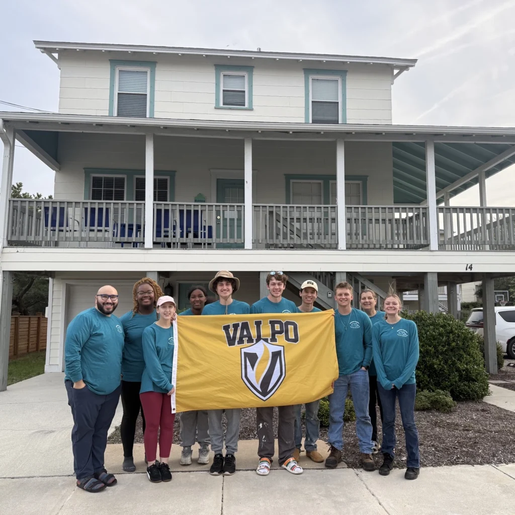 Ten Valparaiso University students in matching teal shirts hold a yellow VALPO flag in front of a white two-story house, celebrating Spring Break 2026 as they serve together.