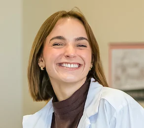 A woman smiling brightly, wearing a white coat and a dark turtleneck sweater, showcases her pride as a participant in the Health Science Program.