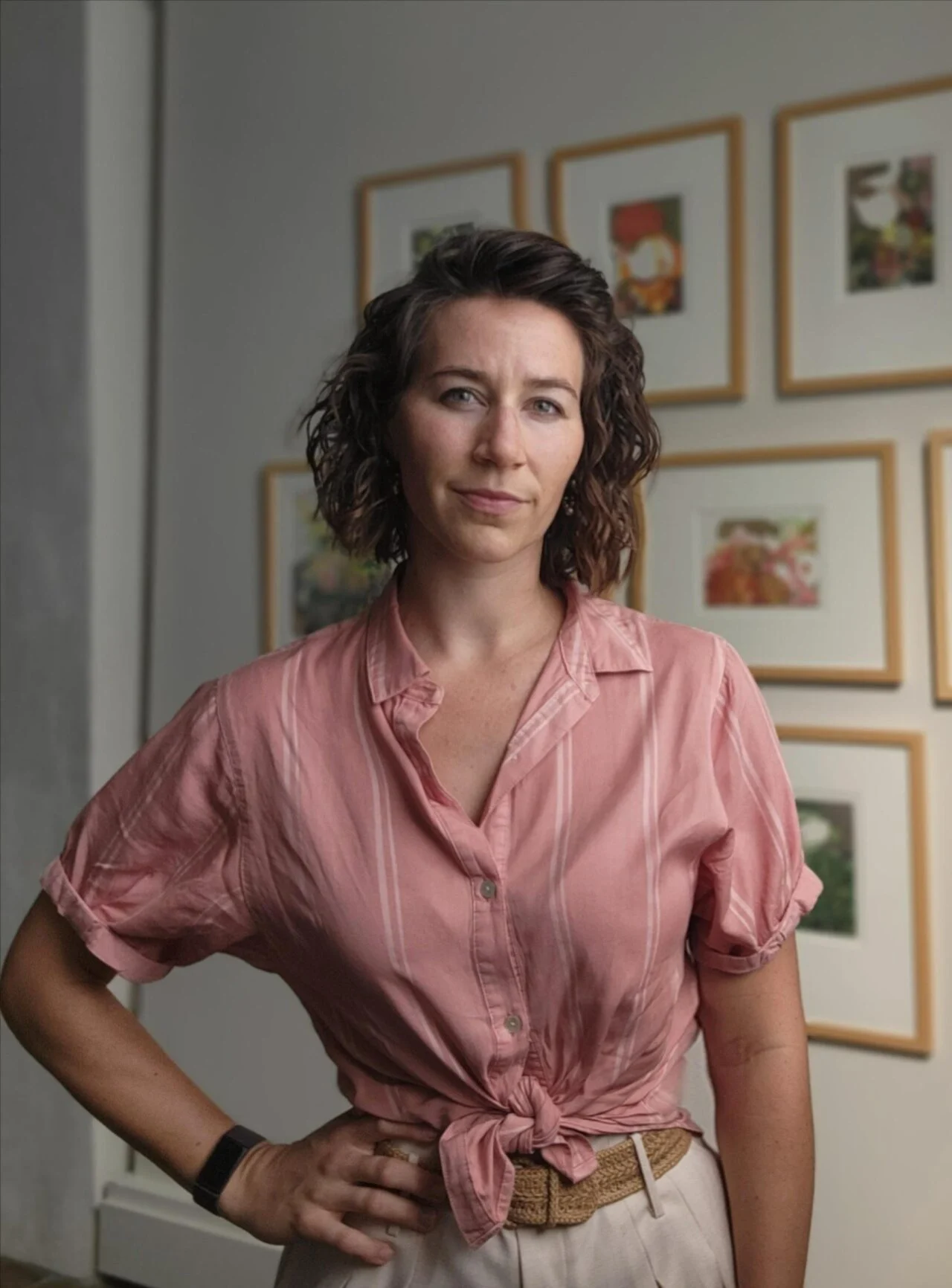 Woman in a pink tied blouse stands confidently indoors before a gallery wall of framed botanical prints, ready to lead inspiring workshops.