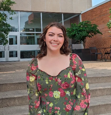 Sarah Dilday, a participant in the Health Science Program, smiles in a green floral dress while standing on steps outside a brick building.