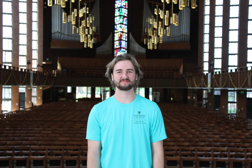 A man in a turquoise shirt stands smiling inside a large, empty church with stained glass and chandeliers at Valparaiso University.