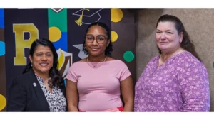 Three women standing together and smiling in front of a colorful graduation-themed backdrop for an OMP Spotlight moment.