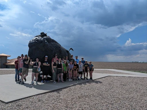 A group of people pose in front of a large buffalo statue on a concrete platform under a cloudy sky, creating a scene straight from a meteorology textbook.