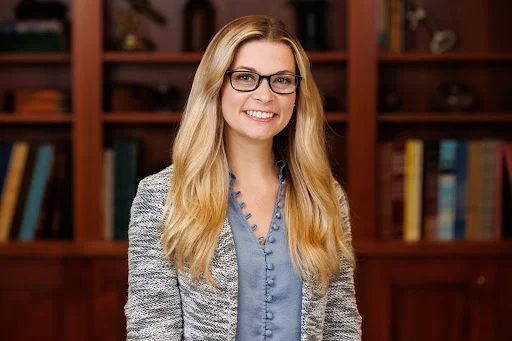 A Valparaiso University faculty member with long blonde hair and glasses smiles while standing in front of bookshelves.
