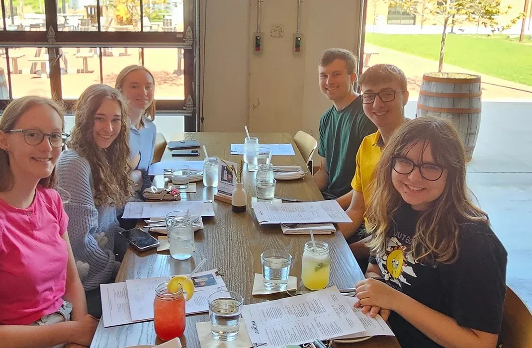 Six young adults sit at a restaurant table with drinks, menus, and smiles, sharing stories about meteorology as they enjoy a meal together.