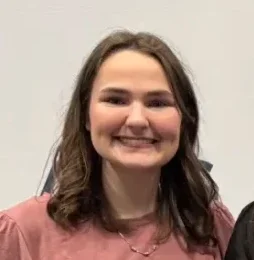 Zoe Barajaz, a woman with shoulder-length brown hair, wears a pink top and smiles in front of a white backdrop with text, reflecting her passion for meteorology.