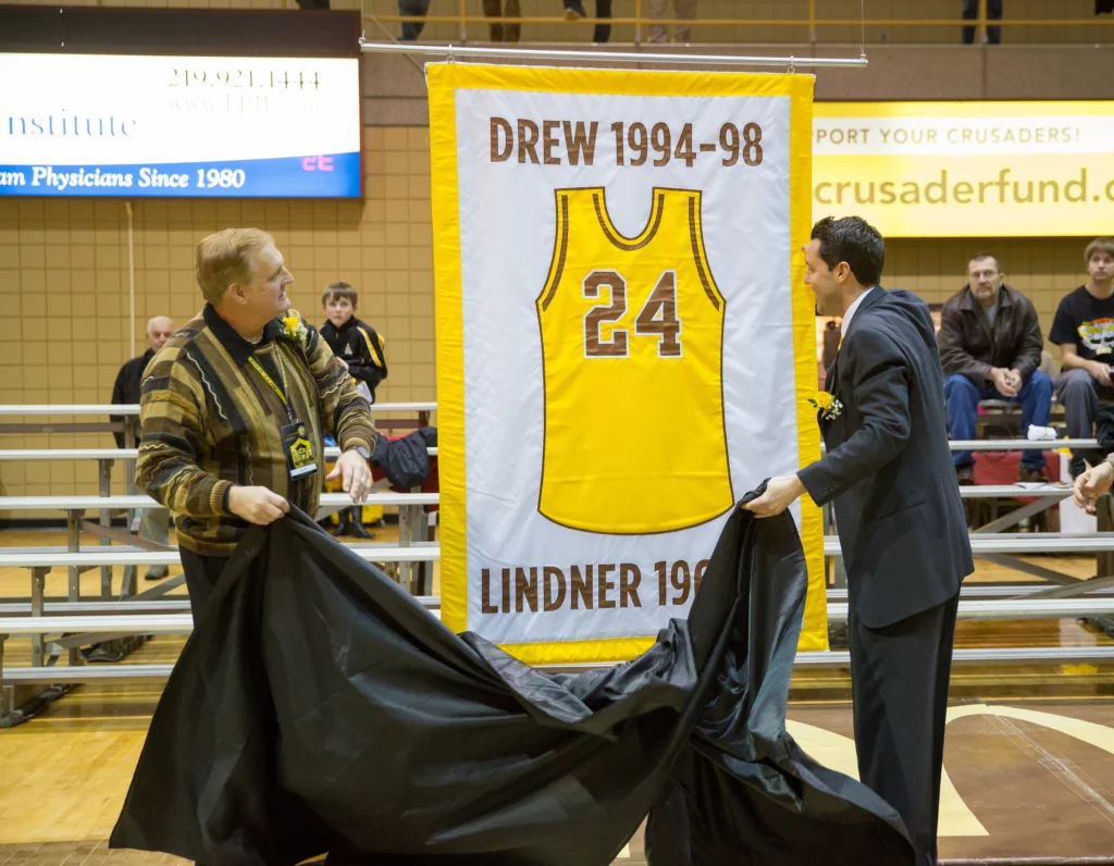 Two men unveil a yellow Valpo basketball jersey banner with the number 24 and the name Drew at a ceremony, honoring Bryce Drew, the 2026 Commencement Speaker.