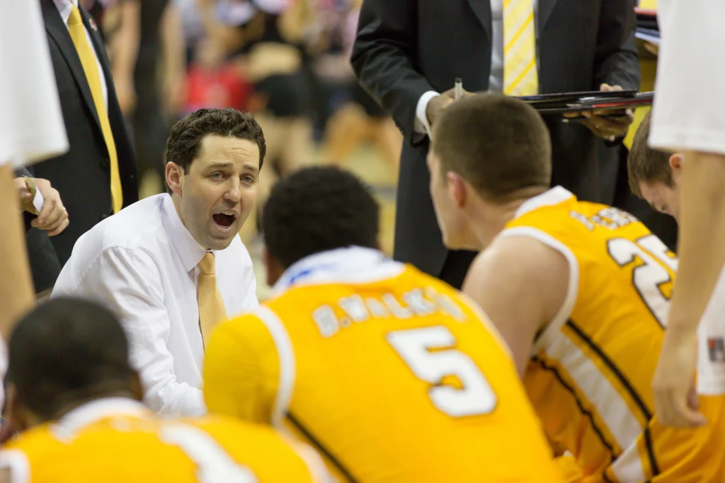 Basketball coach Bryce Drew in a white shirt talks to Valpo players in yellow jerseys during a timeout on the court.