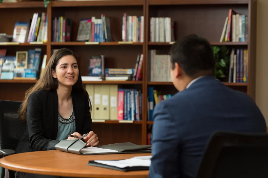 Two people sit at a round table in an office, having a conversation about Organizational Leadership in front of bookshelves.