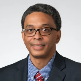 Michael Chikeleze, wearing glasses, a dark suit, blue striped shirt, and red tie, smiles against a plain background.