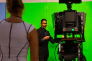 A man in a suit presents in front of a green screen, discussing interesting majors, while a woman and camera are in the foreground.