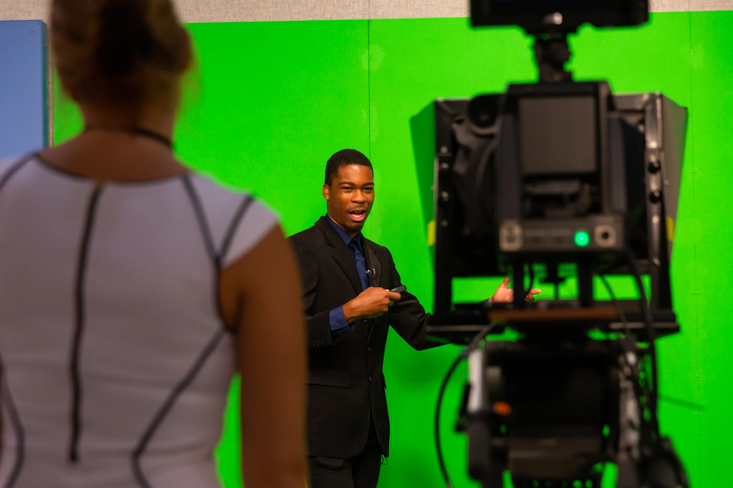 A man in a suit presents in front of a green screen, discussing interesting majors, while a woman and camera are in the foreground.