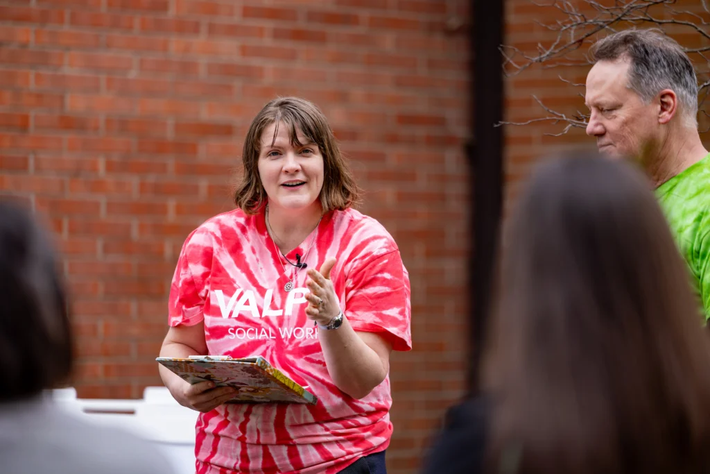 Woman in a red tie-dye shirt speaks to a small group outside, holding a clipboard in front of a brick wall—part of Valparaiso University's public safety social work certificate program.