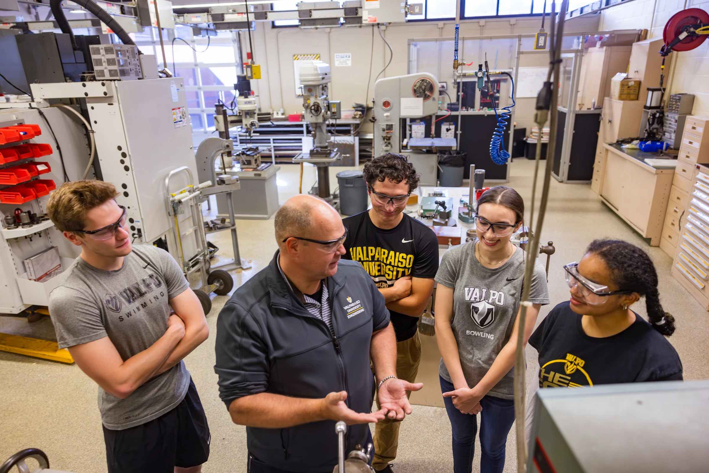 Five people in safety glasses stand in a workshop, attentively listening to an instructor explain equipment as part of an Integrated Business and Engineering course.