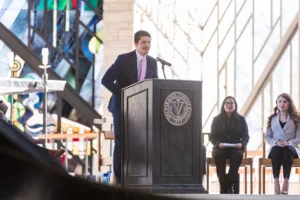 A man in a suit speaks at a podium indoors, with two women seated behind him near stained glass windows.