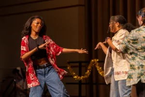 Two people in colorful shirts and jeans dance together on stage with gold tinsel in the background, their movements inspired by Taiya Girley’s signature flair. The vibrant scene glimmers, reminiscent of Beacon’s Reflection shining through each joyful step.
