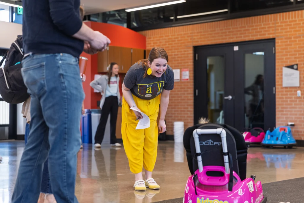 A woman in yellow pants smiles and bends toward a mobility-challenged child in a toy car indoors, highlighting accessibility and care at Valparaiso University.