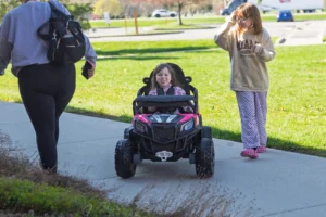 A young girl drives a pink toy car on a sidewalk, showcasing accessibility for mobility-challenged children, while another girl and an adult walk nearby on a sunny day.