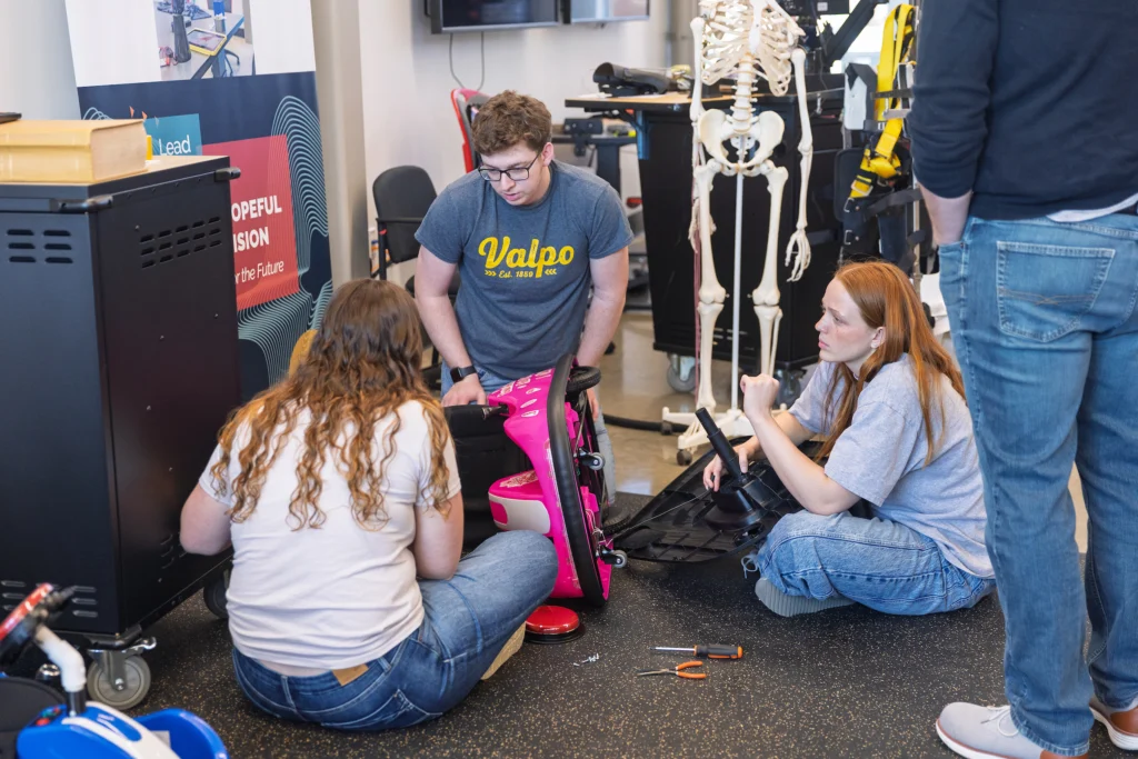 Three people sit on the floor assembling a pink wheelchair, surrounded by tools and equipment, working together to improve accessibility for mobility-challenged children.