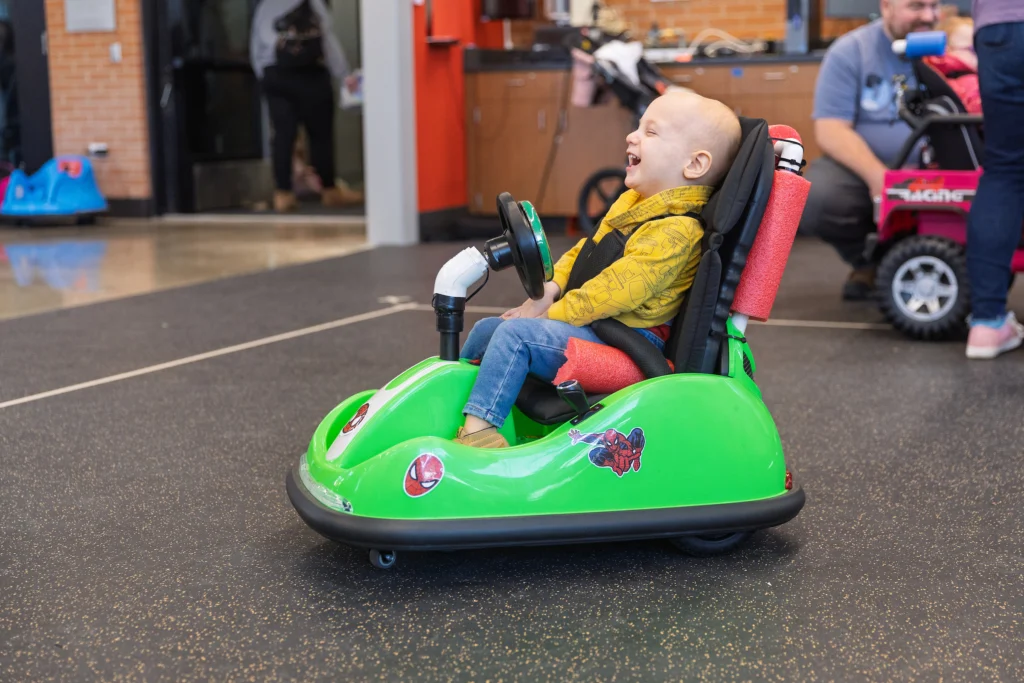 A young child joyfully rides a small green toy car indoors, smiling and laughing—a moment highlighting the importance of accessibility for mobility-challenged children.