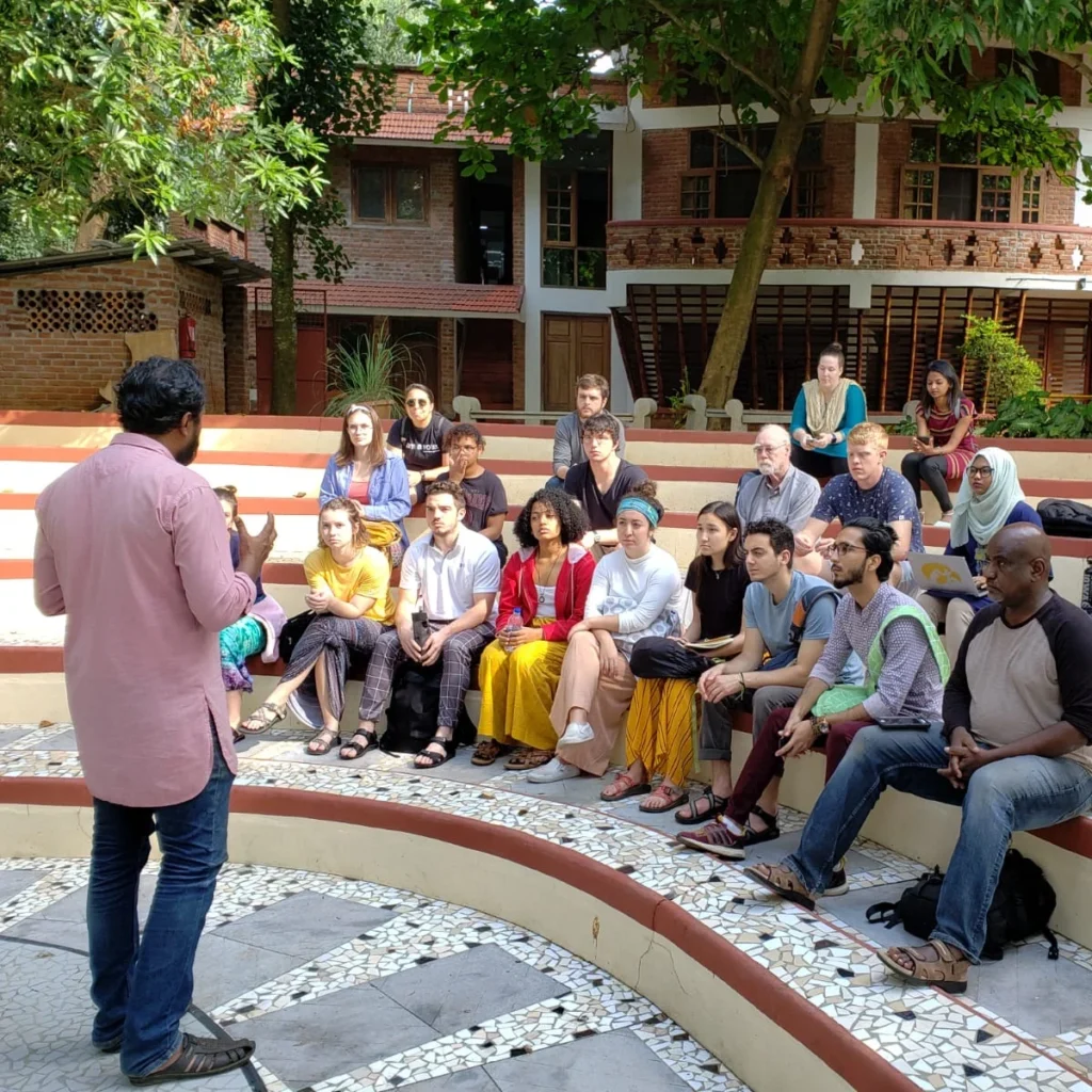 A group of people sit outdoors on tiered steps, listening to plenaries delivered by a speaker in front of trees and brick buildings.
