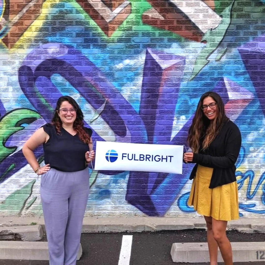 Two women hold a Fulbright sign in front of a colorful graffiti wall in a parking lot, celebrating after attending inspiring plenaries.