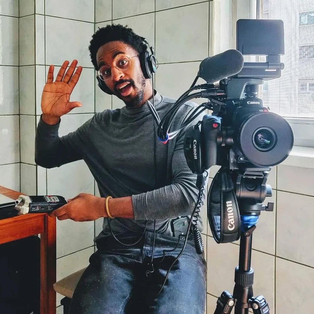Man wearing headphones waves and smiles at the camera while sitting next to video equipment, a Canon camera, and prepares for plenaries.