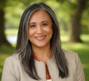 Smiling woman with long gray hair stands outdoors, wearing a beige jacket and red necklace—Dr. Mary Grace Almandrez, Senior Vice President at Valparaiso University.