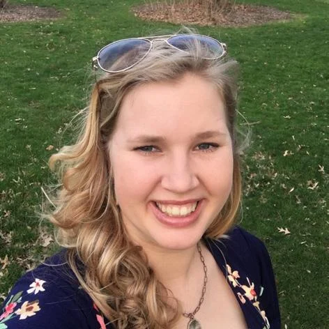 Smiling woman, Olivia VanTol, with long blonde hair and sunglasses on her head, standing on grass outdoors at IBE.