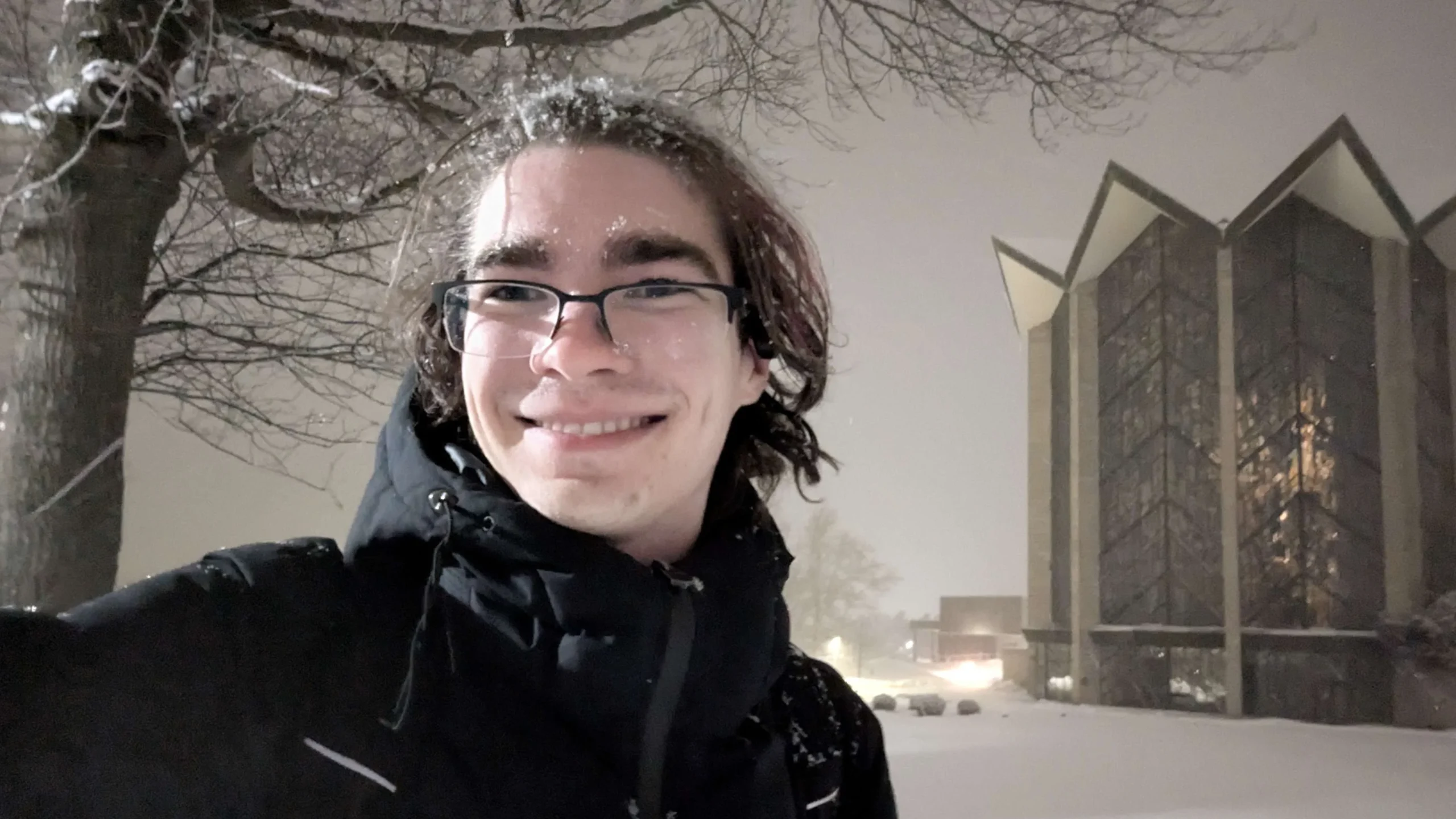 Person with glasses and dark jacket taking a selfie outside in the snow at night near a modern building on the Valpo campus, capturing a moment in the vibrant community.