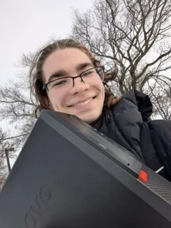 Person with glasses smiling outdoors at Valpo, holding a black Lenovo computer, surrounded by bare trees—a proud member of the Computer Engineering student community.