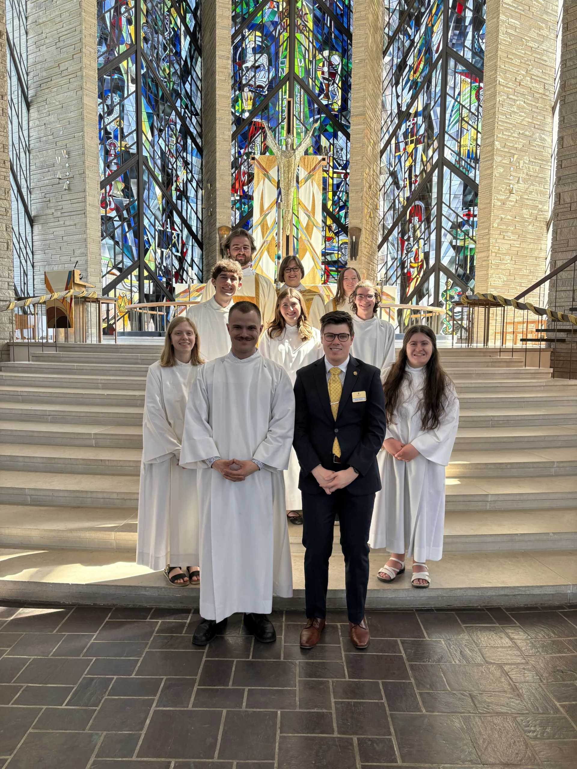 Nine members of the Valpo student community in white robes and one in a suit pose on church steps in front of colorful stained glass windows, celebrating achievements in fields like Computer Engineering.