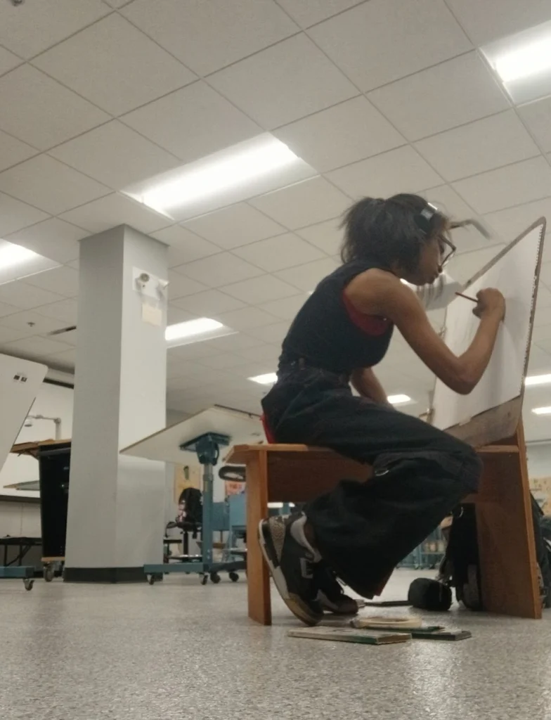 A person sits on a bench, sketching on a large drawing board in an art classroom, capturing Beacon's Reflection with inspiration drawn from Taiya Girley.