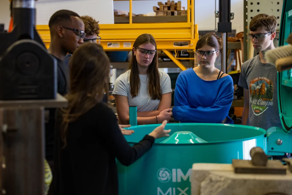 A group of students wearing safety glasses gather around a large teal machine in a workshop, exploring interesting majors that lead to hands-on learning and surprising careers.