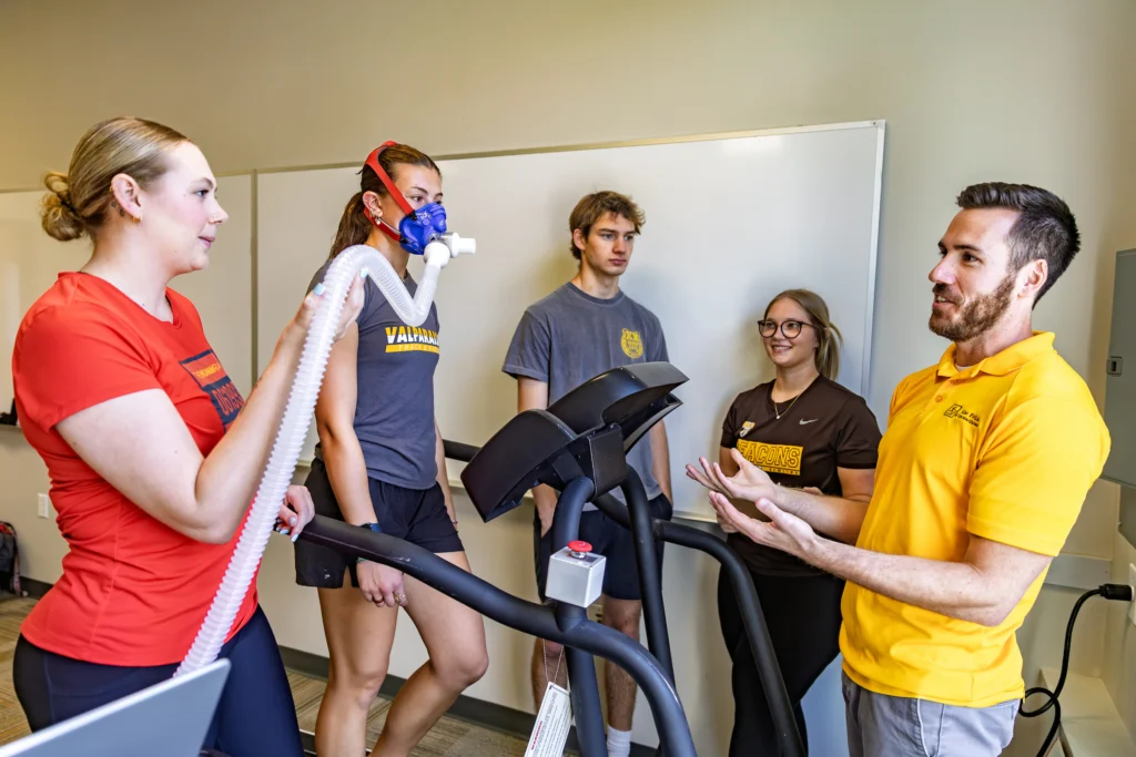 Four students and an instructor gather around treadmills as one student wears a fitness mask for a test, showcasing the hands-on experience found in interesting majors related to health and exercise careers.