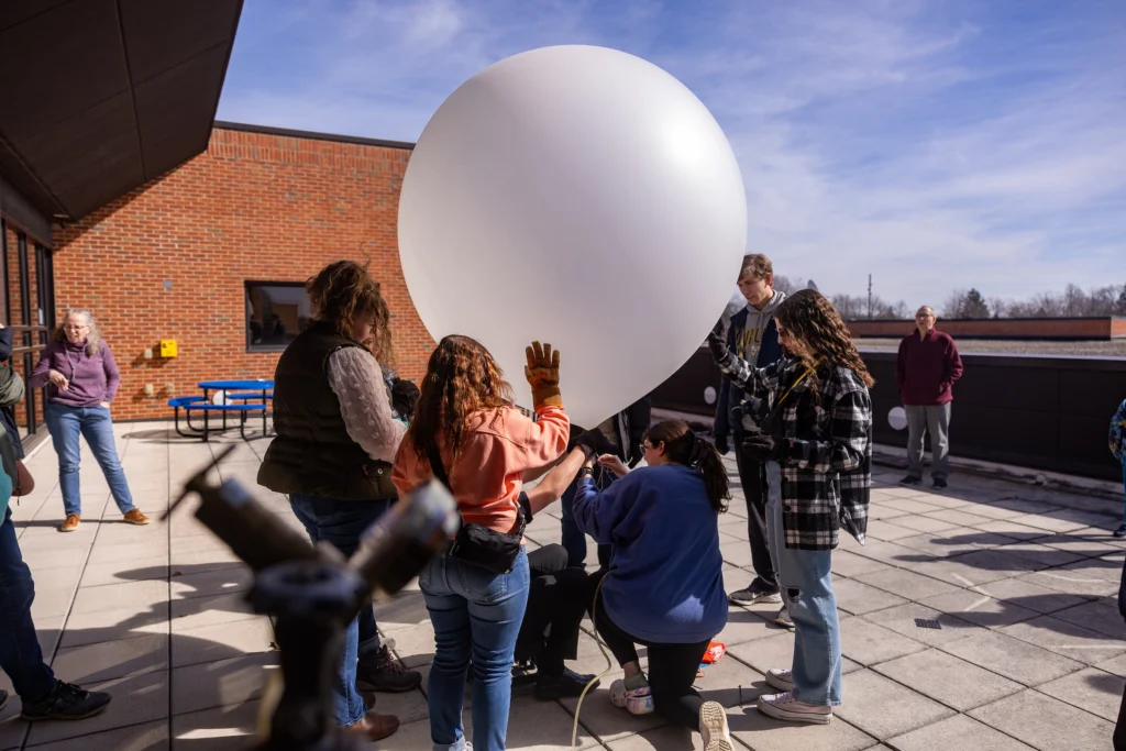 A group of people gather on a rooftop, inflating a large white weather balloon under a blue sky—perhaps exploring interesting majors or surprising careers in atmospheric science.