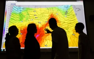 Four people at Valparaiso University examine a colorful weather map projection showing wind patterns, temperature gradients, and notable ozone spikes.
