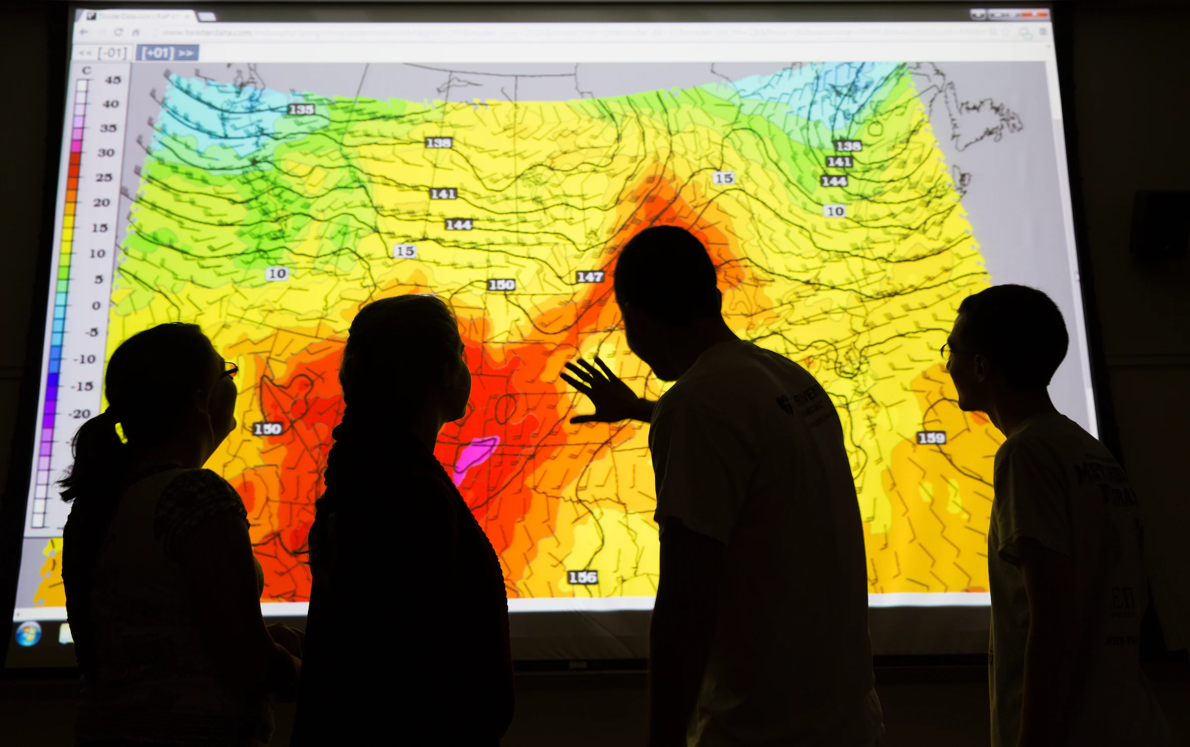 Four people at Valparaiso University examine a colorful weather map projection showing wind patterns, temperature gradients, and notable ozone spikes.