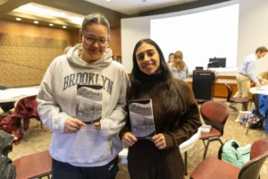 Two Valpo students smiling and holding their copies of Letras, standing in a room with people and tables in the background.
