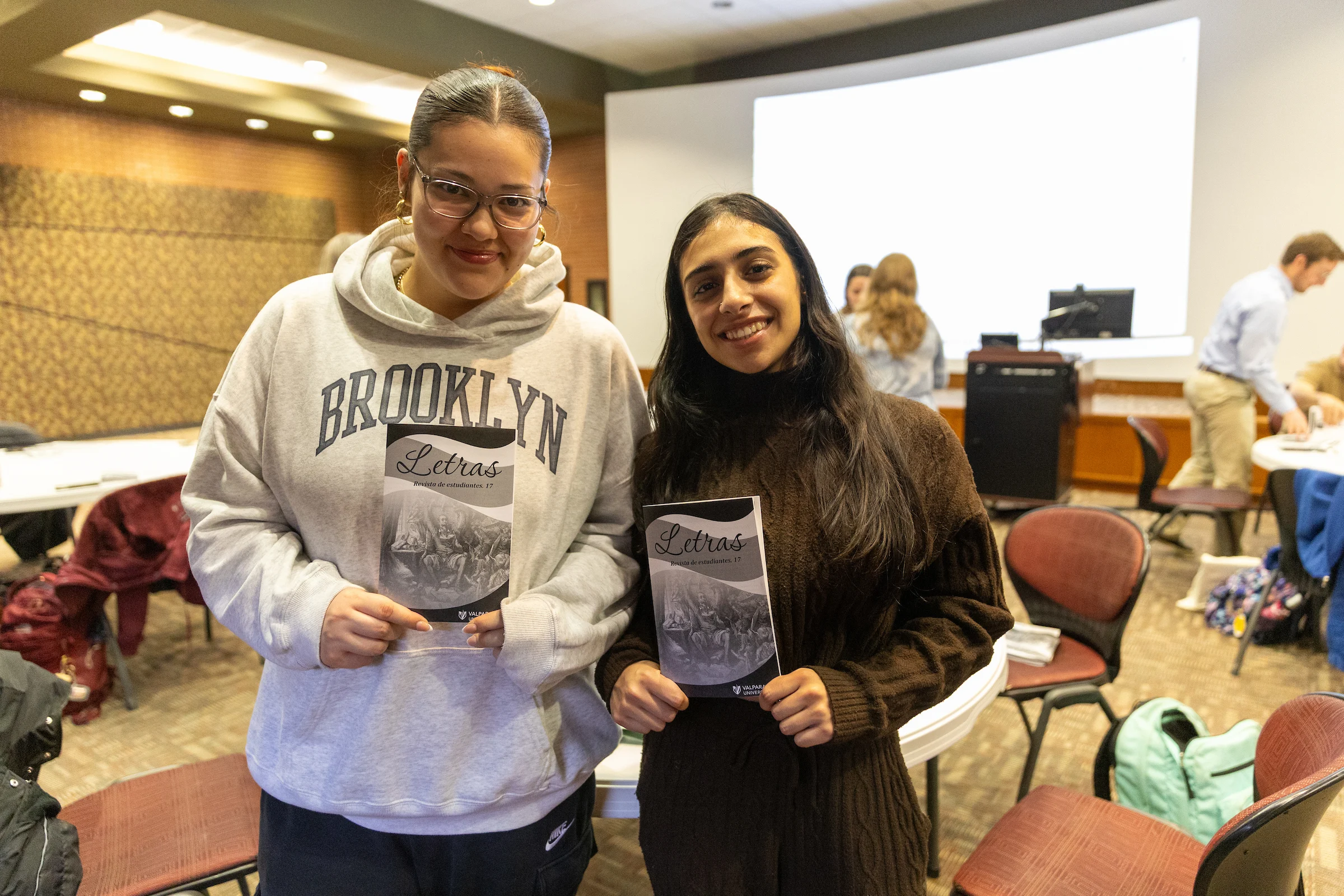 Two Valpo students smiling and holding their copies of Letras, standing in a room with people and tables in the background.