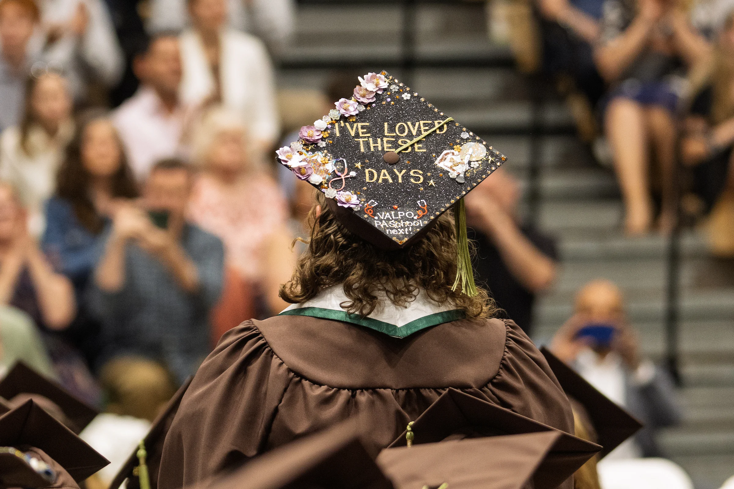 Graduate in cap and gown with decorated cap reading "I'VE LOVED THESE DAYS" at a ceremony, reflecting lessons and appreciation, with the crowd in the background.