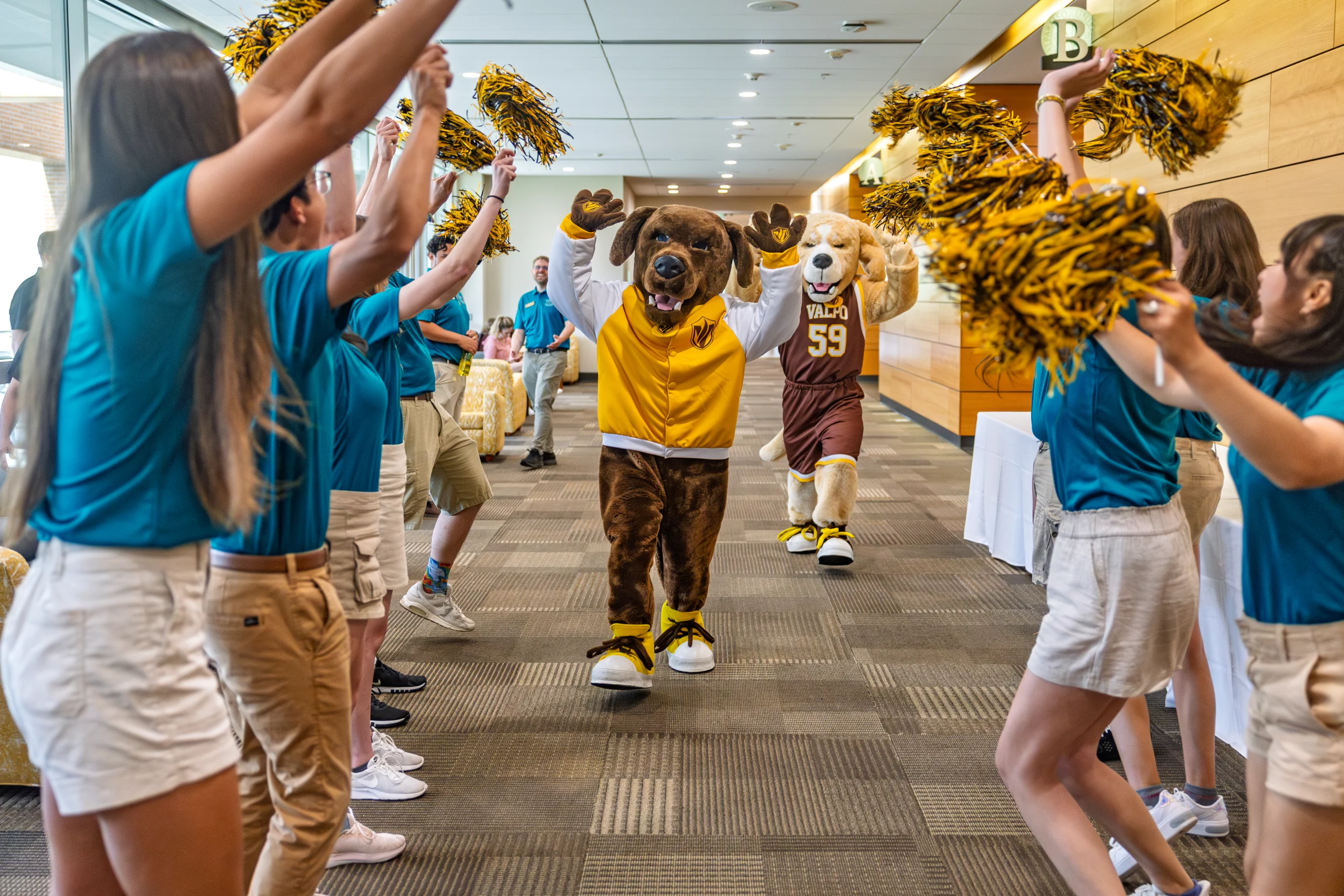 Two animal mascots walk through a tunnel of cheering people holding yellow and black pom-poms indoors, creating an energetic atmosphere that celebrates Student Involvement.