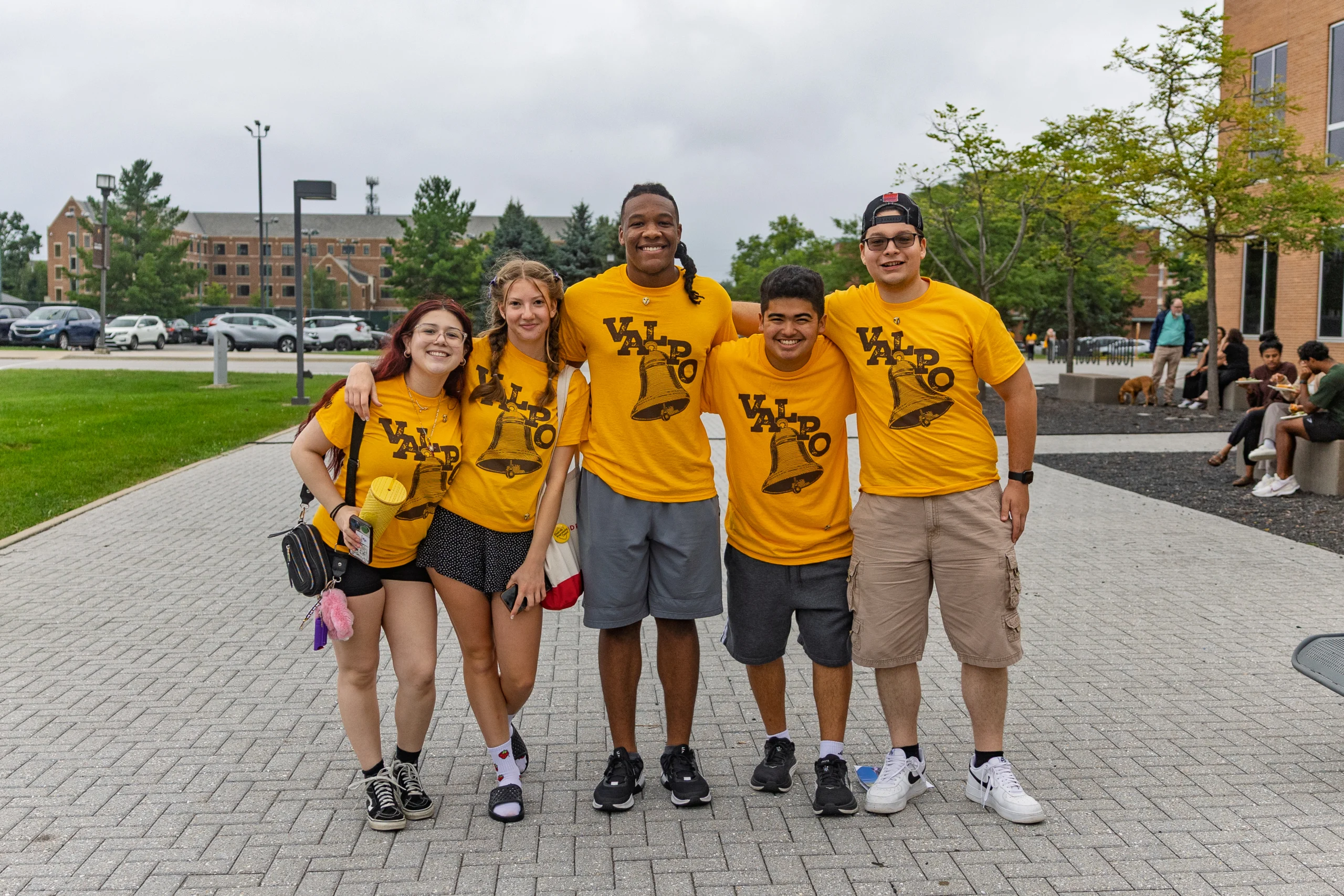 Five smiling students in matching yellow t-shirts, showcasing student involvement, pose together outdoors on a college campus walkway.