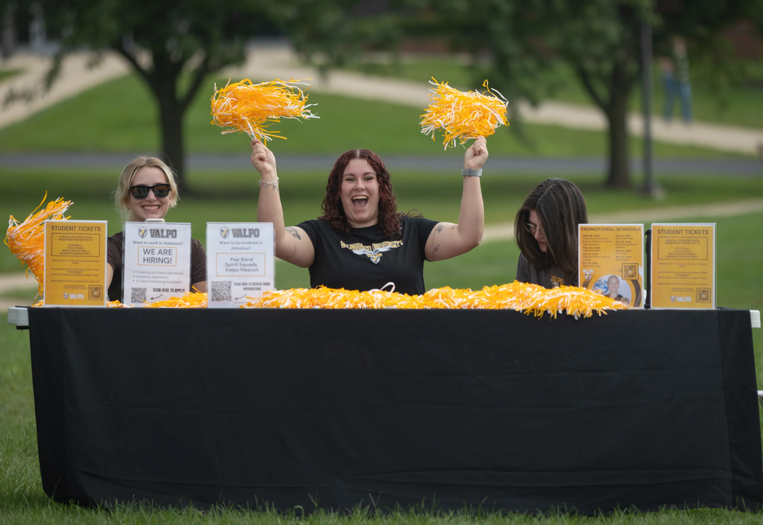 Three people at an outdoor table with yellow pom-poms, smiling and handing out student tickets to promote Student Involvement.