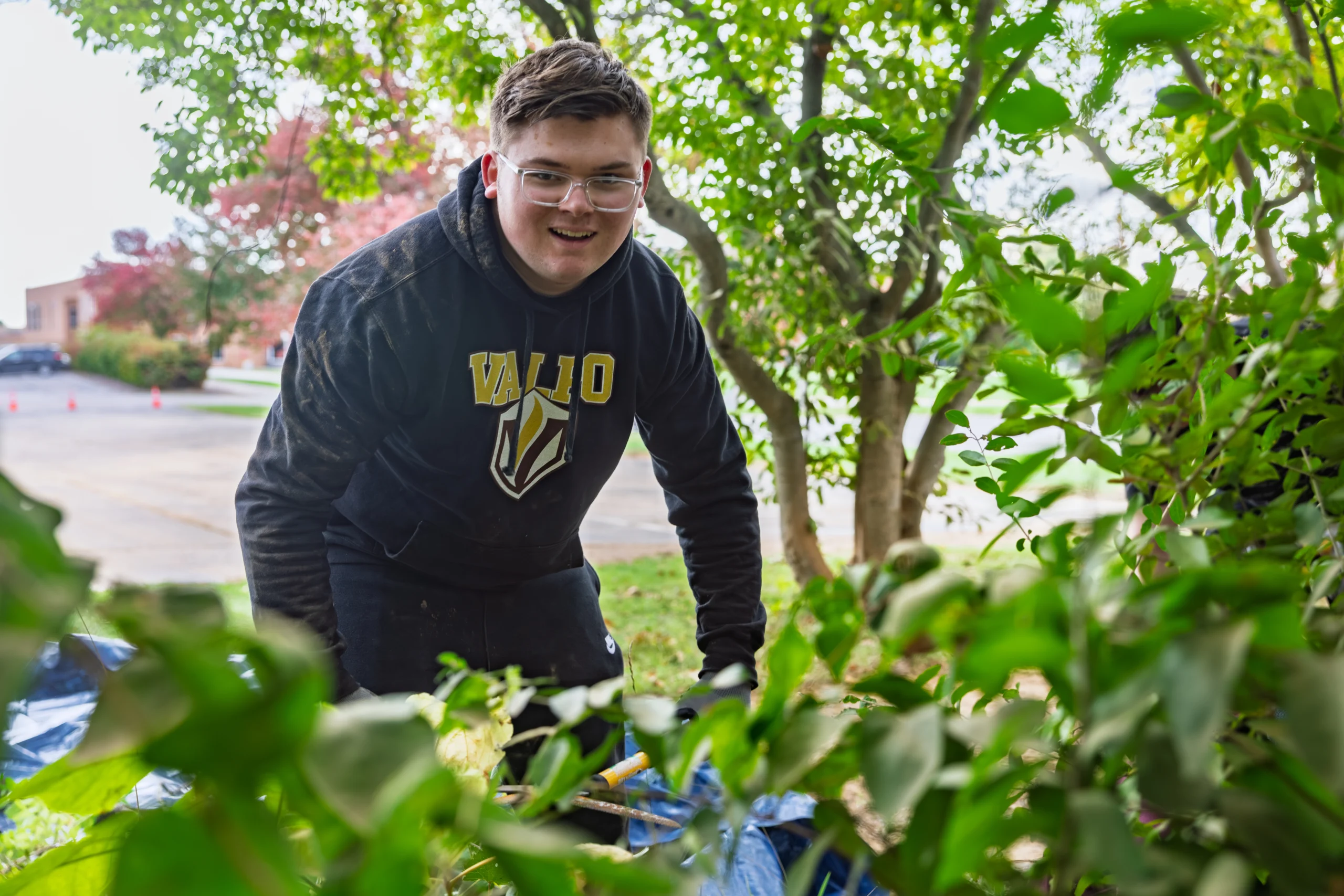 A person in glasses and a black hoodie works in a garden, smiling among green plants and trees, showcasing the benefits of student involvement in outdoor activities.