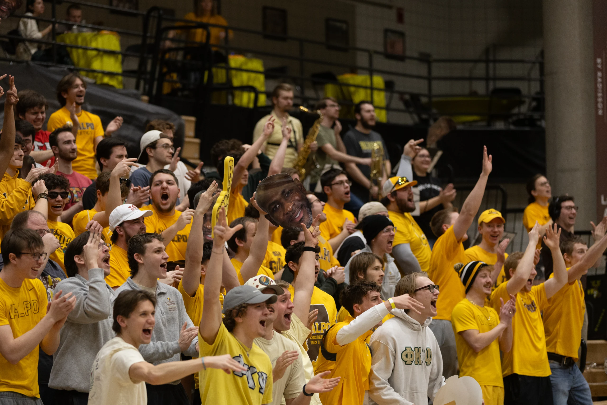 Crowd of cheering students in yellow shirts at a sports event, showcasing strong student involvement, with some holding signs and cutouts.